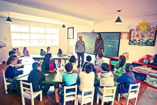 Children at long green tables in a classroom, two adults standing by the blackboard. Bogotá, Colombia, 2011.