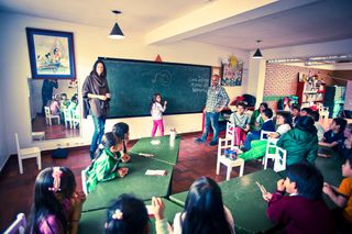 A classroom seen from the back: children at green tables, a woman and a man at the blackboard. Bogotá, Colombia, 2011.