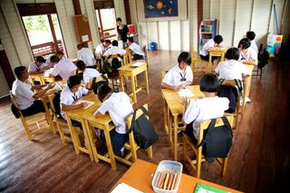 A classroom of students in white uniforms drawing at wooden desks. Thailand, 2011.
