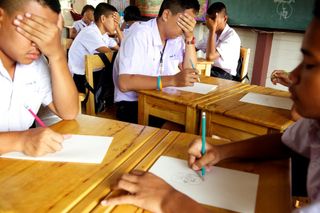 Students drawing at wooden desks, one covering his eyes with his hand. Thailand, 2011.