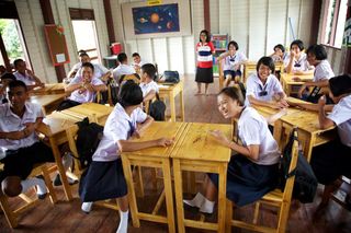Students at wooden desks in a classroom, a teacher standing at the back. Thailand, 2011.