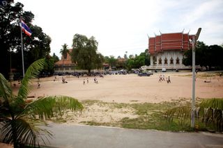 A wide sandy schoolyard with children playing, a temple roof beyond the trees. Thailand, 2011.