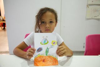 A girl at a white table holds up her drawing of a green alien on orange ground. Barcelona, Spain, 2010.
