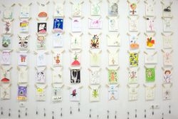 A white wall hung with rows of small children's drawings on wooden pegs. Barcelona, Spain, 2010.
