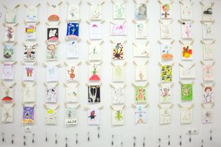 A white wall hung with rows of small children's drawings on wooden pegs. Barcelona, Spain, 2010.