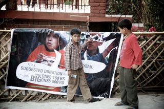 Two boys stand beside a large printed banner propped against a bamboo fence. Rishikesh, India, 2010.