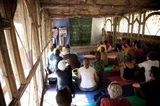 People sit on mats in a long bamboo hall facing a green hanging cloth. Rishikesh, India, 2010.
