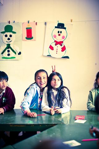 Two girls smiling at a green table, paper snowmen pinned to the wall behind them. Bogotá, Colombia, 2011.