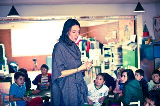 A woman in a grey shawl talking to children seated at their tables. Bogotá, Colombia, 2011.