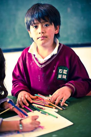 A boy in a maroon school sweater sits behind a pile of coloured pencils. Bogotá, Colombia, 2011.