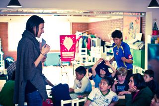 A woman in a grey shawl stands among seated children, a pink poster on the wall. Bogotá, Colombia, 2011.