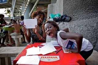 Two girls laughing at a red table covered in paper, one holding up a sheet. Brazil, 2012.
