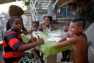 Children drawing around a green table on a terrace, one boy in a striped shirt. Brazil, 2012.