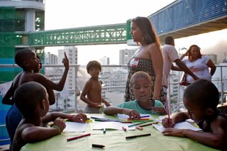 Children drawing at a green table on a terrace, a woman standing behind them. Brazil, 2012.