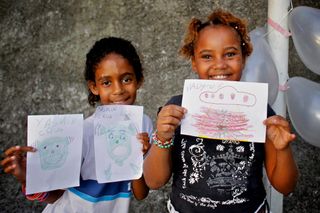 Two girls hold up their alien drawings in front of a concrete wall. Brazil, 2012.