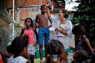 A woman in white talks with a group of children outside among brick houses. Brazil, 2012.