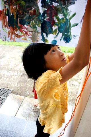 A girl in a yellow top reaches up to a painted sheet hanging on a wall. Indonesia, 2011.