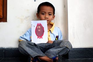 A boy sits cross-legged against a wall, holding a drawing of a red egg-shaped alien. Indonesia, 2011.