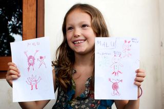 A smiling girl holds up two sheets of small pink alien drawings. Indonesia, 2011.