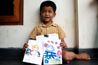 A boy sits against a wall with two colourful alien drawings on his knees. Indonesia, 2011.
