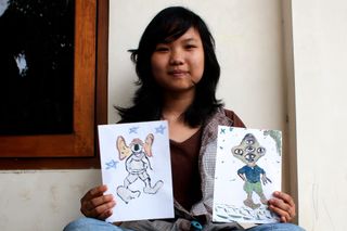 A young woman holds up two drawings of aliens with big eyes. Indonesia, 2011.