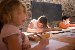 Children drawing at wooden tables in a room with stone walls and a blackboard. Mexico, 2011.
