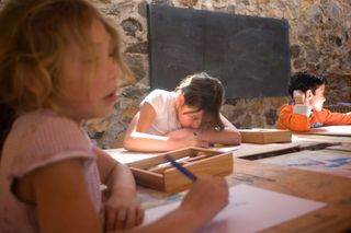 Children drawing at wooden tables in a room with stone walls and a blackboard. Mexico, 2011.