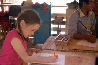 A girl in pink draws with an orange pencil at a wooden table. Mexico, 2011.