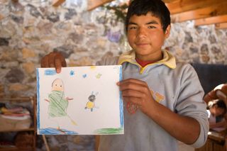 A boy holds up a drawing of a green figure and a small creature in the sky. Mexico, 2011.
