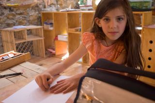A girl in an orange top sits at a wooden table with a blank sheet. Mexico, 2011.