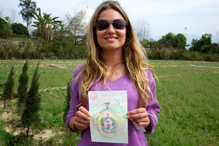 A woman in sunglasses holds up a small round drawing on a lawn. Rishikesh, India, 2012.