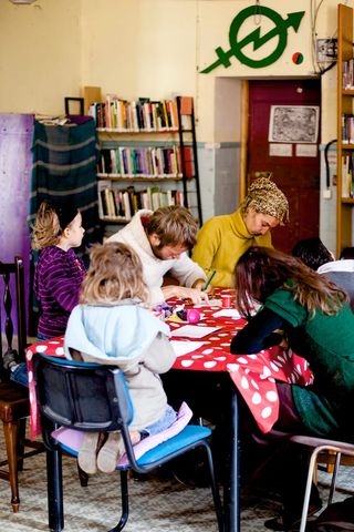 Children and adults drawing at a table with a red spotted cloth in a room with bookshelves. Spain, 2012.
