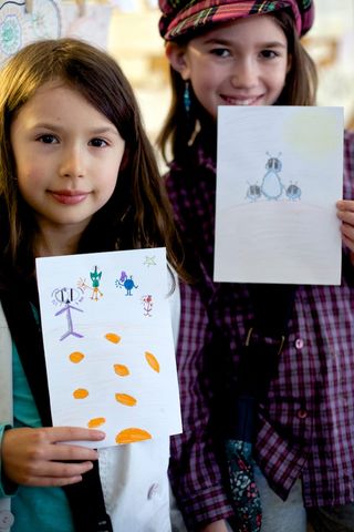 Two girls hold up their drawings, one with orange dots and small figures. Spain, 2012.