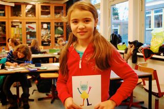 A girl in a red jacket holds an alien drawing with smiling green face and stick limbs among other children at art tables. Germany, 2011.