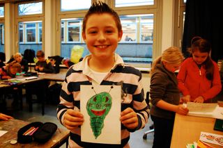 A boy in a black-and-white striped shirt holds a green alien drawing with antenna, classroom in background. Germany, 2011.