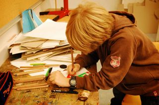 A girl in a red shirt concentrates on drawing at a shared table with paint palettes and brushes. Germany, 2011.