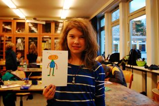 A girl in a navy-striped shirt holds an orange and pink alien drawing with round features. Germany, 2011.