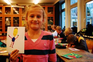 A girl in pink and navy striped sweater holds a yellow-faced alien drawing with outstretched arms. Germany, 2011.