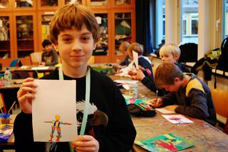 A boy holds a colourful stick-figure alien drawing with extended limbs and bright paint colours. Germany, 2011.