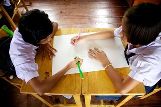 Two students draw on one big sheet from opposite sides of a wooden table. Thailand, 2011.