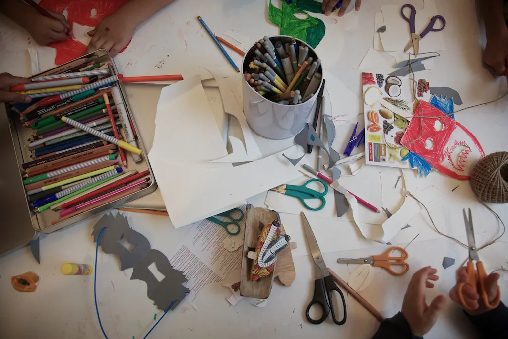 A worktable seen from above, covered with pens, scissors, paper cut-outs and a tin of pencils.