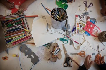 A worktable seen from above, covered with pens, scissors, paper cut-outs and a tin of pencils.