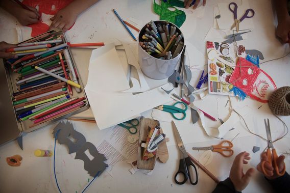 A worktable seen from above, covered with pens, scissors, paper cut-outs and a tin of pencils.