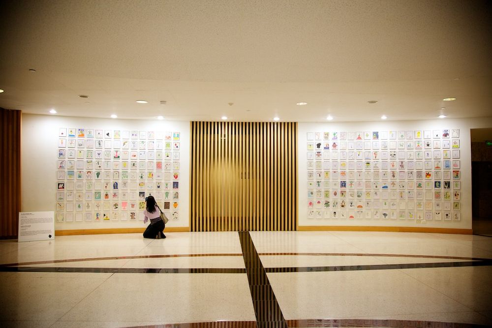 Two long walls of children's drawings hung in rows in the Bangkok Art and Culture Centre, with one visitor kneeling in front of them.