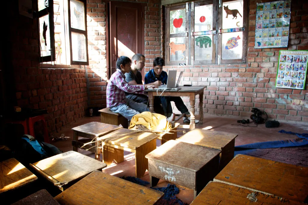 Adults and a young person working at a laptop in a room with wooden desks.
