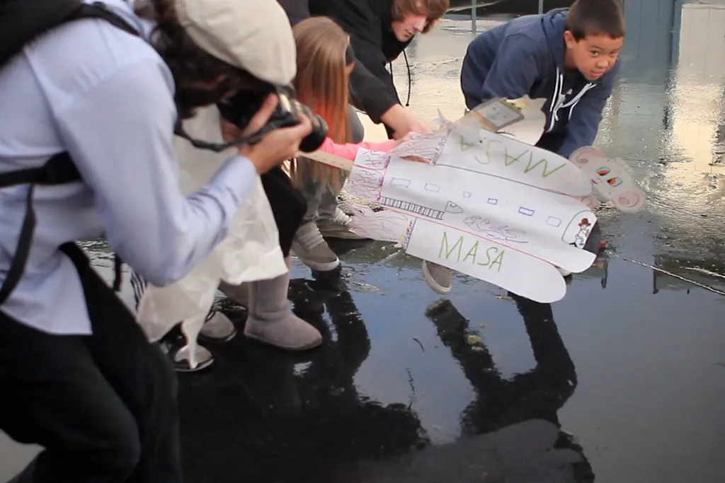 Teenagers photographing paper cut-outs laid on a reflective floor.