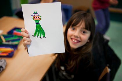 A child holding up a drawing of a green alien at a table.