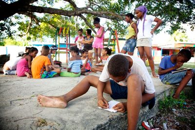 Children drawing outdoors on the ground under a large tree.