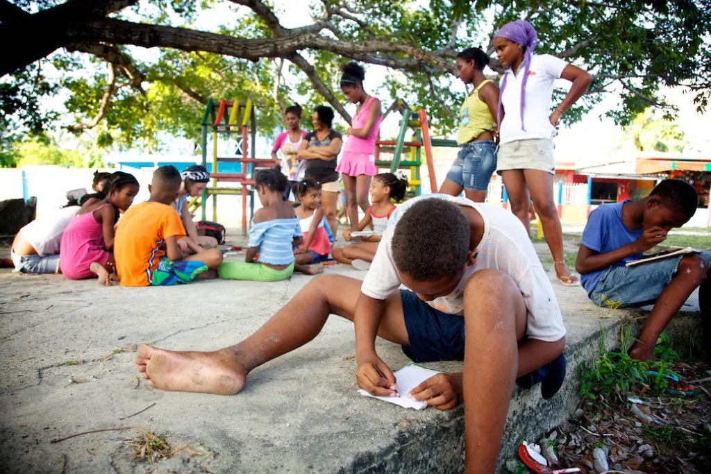 Children drawing outdoors on the ground under a large tree.