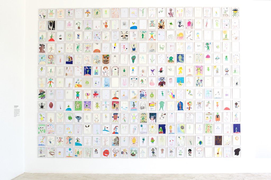 A white gallery wall hung with hundreds of alien drawings in a tight grid.